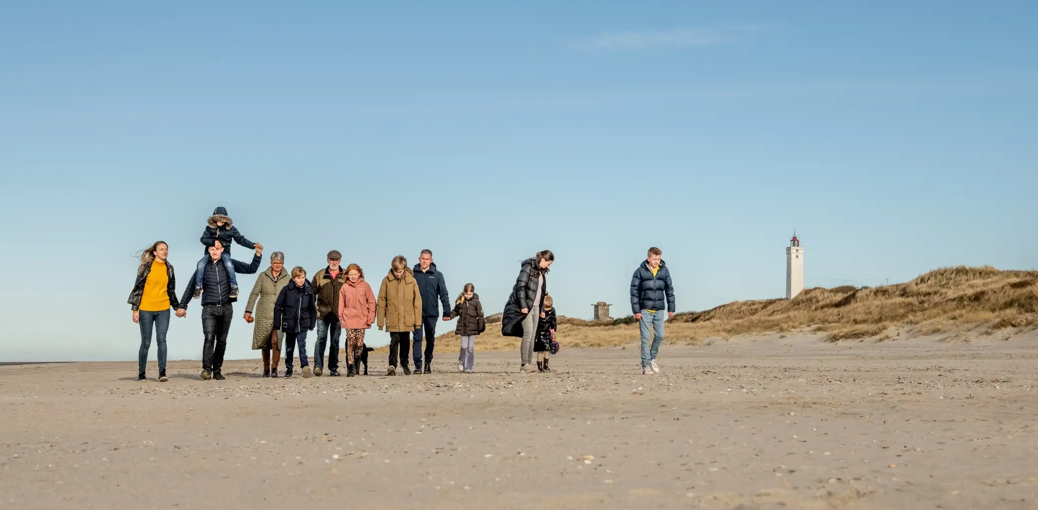 Familie går på stranden med klitter og fyr i baggrunden