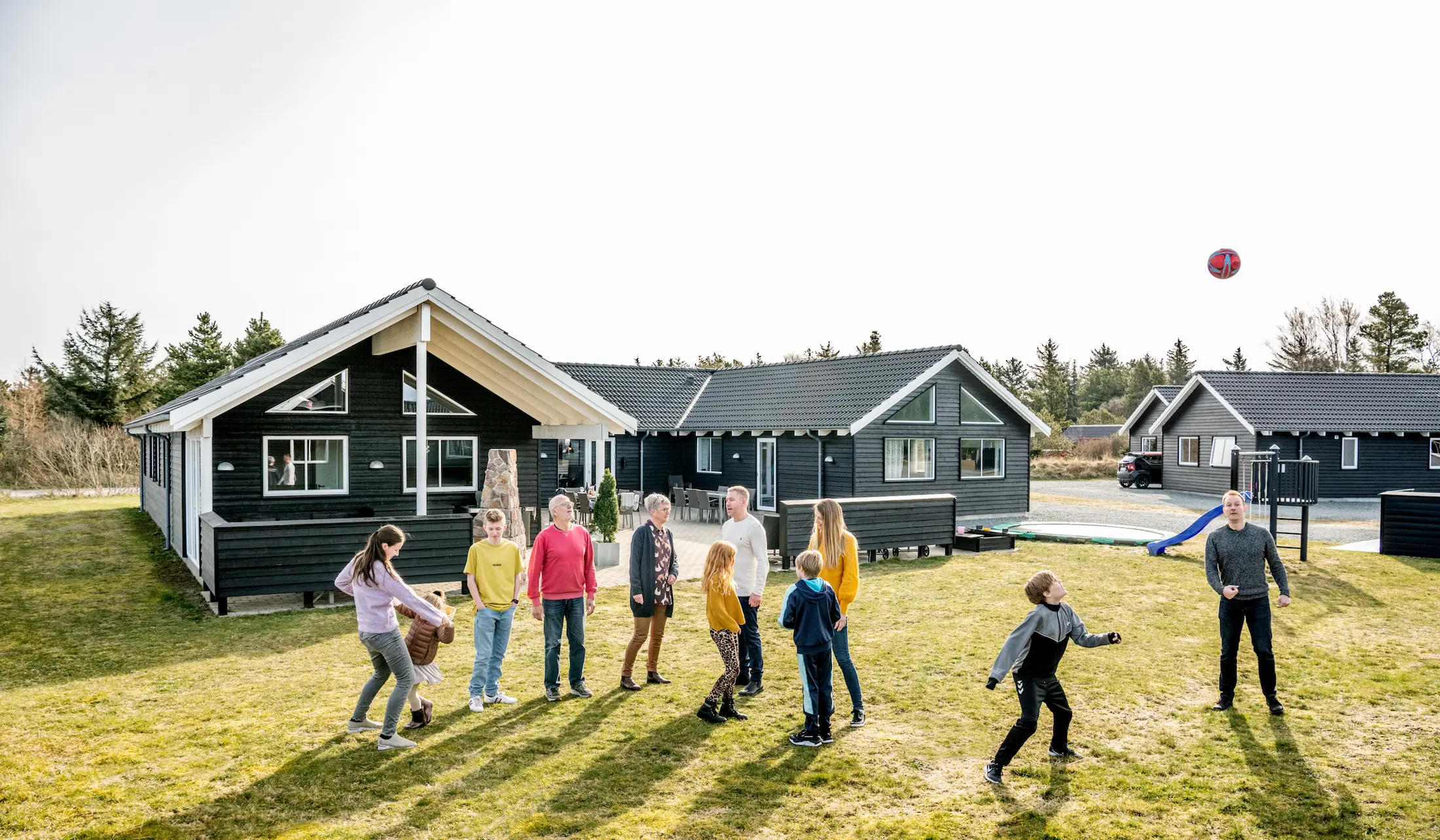 Børn og voksne står på græsplæne udenfor sommerhus og er aktive med hinanden og fodbold